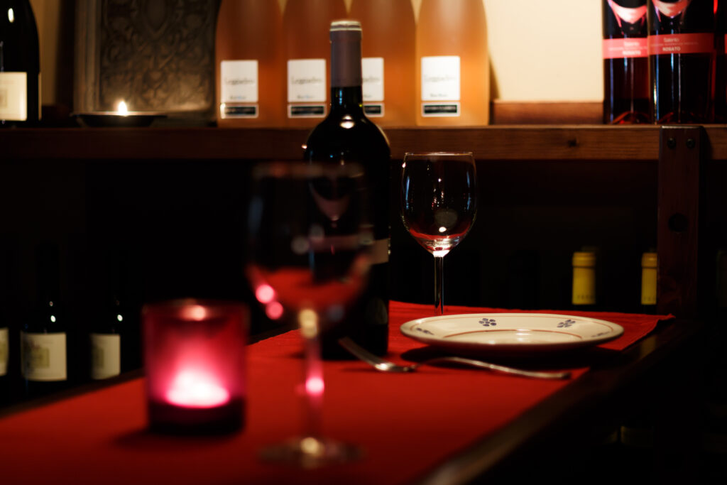 Un tavolo apparecchiato per la cena, scarsamente illuminato, con una candela, una bottiglia di vino, un bicchiere e un piatto su una tovaglia rossa. Bottiglie di vino riempiono gli scaffali sullo sfondo.