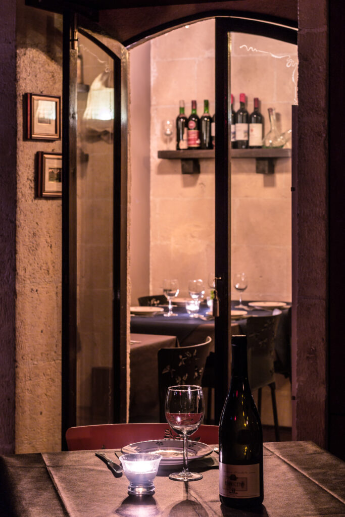Accogliente ristorante a Gioia del Colle, Vineria Rocca Cellarii, con luci soffuse; un tavolo illuminato da candele ospita una bottiglia di vino e un bicchiere, il tutto ammirato attraverso una finestra che mostra un interno arredato in modo accogliente.