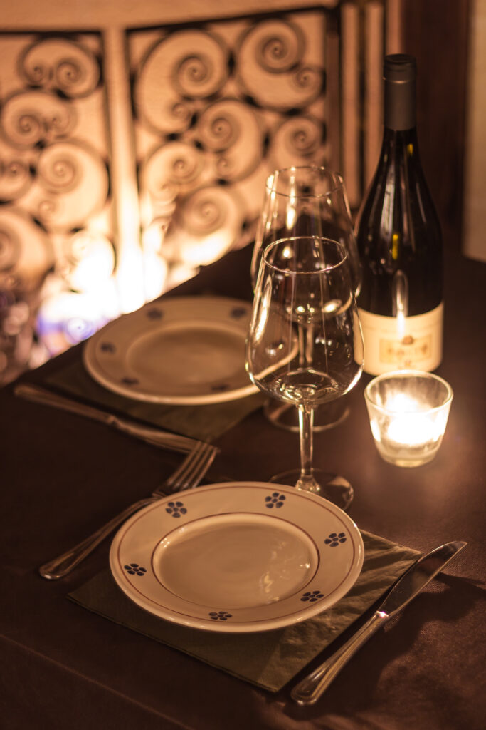 Elegante allestimento da pranzo con due piatti, bicchieri da vino e una bottiglia accanto a una candela accesa. Atmosfera calda e intima, impreziosita da raffinati elementi in metallo, in Vineria Rocca Cellarii.