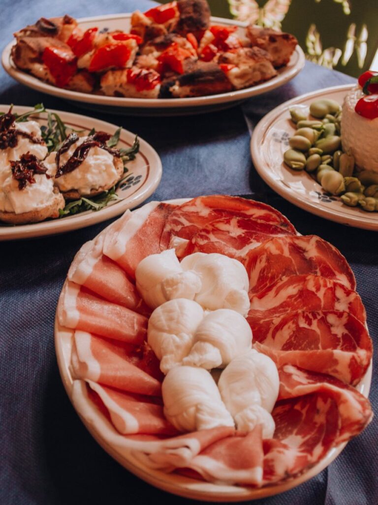 In primo piano un piatto di antipasti con prosciutto, capocollo e mozzarella locale, circondato da piatti di bruschette, formaggi e fave su una tovaglia scura.
