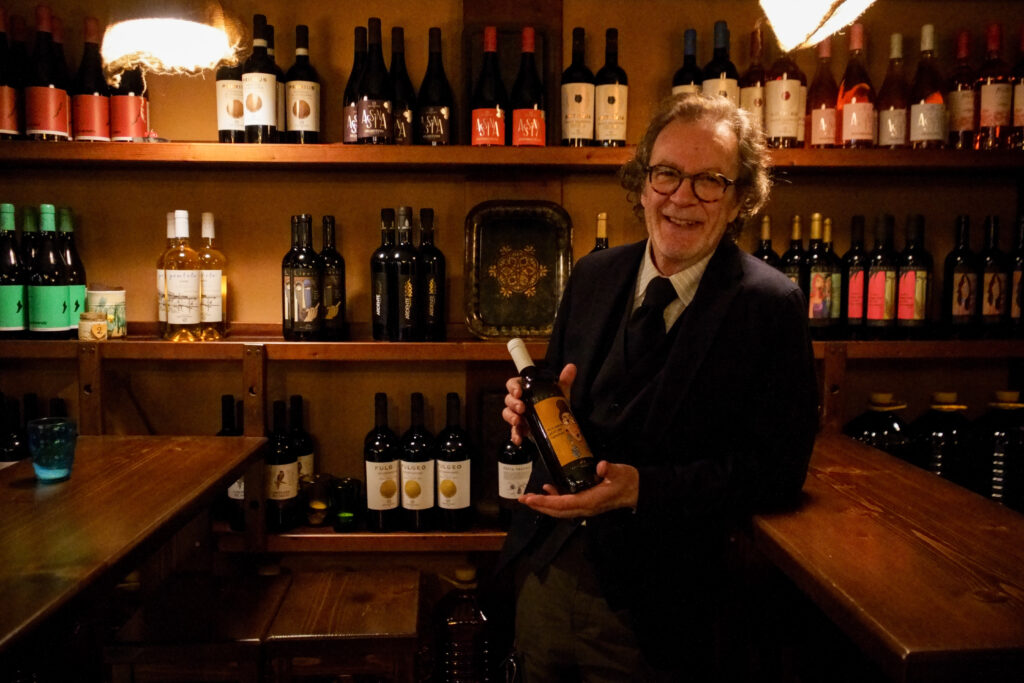 Un uomo in giacca e cravatta sorride calorosamente, tenendo in mano una bottiglia di vino in una cantina accogliente e scarsamente illuminata, piena di bottiglie di vino assortite su scaffali di legno: è la Vineria Rocca Cellarii a Gioia del Colle