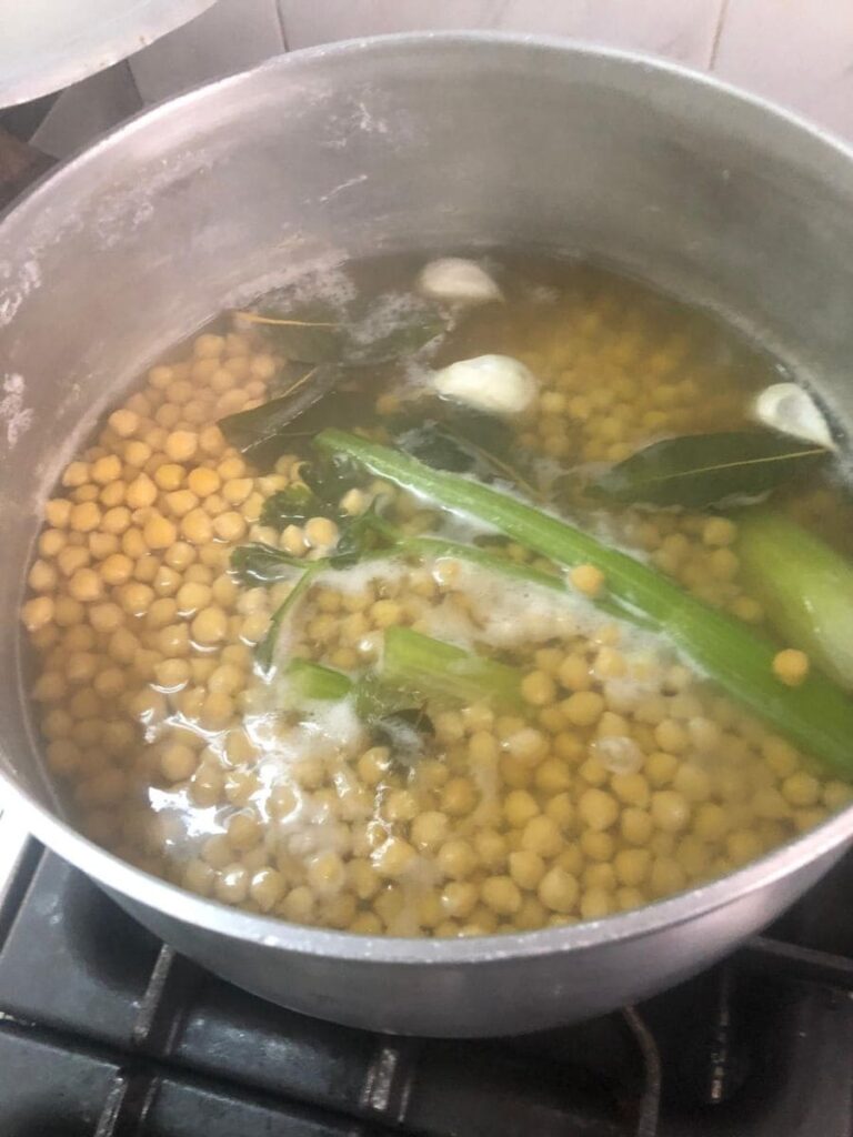 Una pentola con ceci in lenta cottura, insieme a sedano e aromi freschi, durante la preparazione della ricetta.